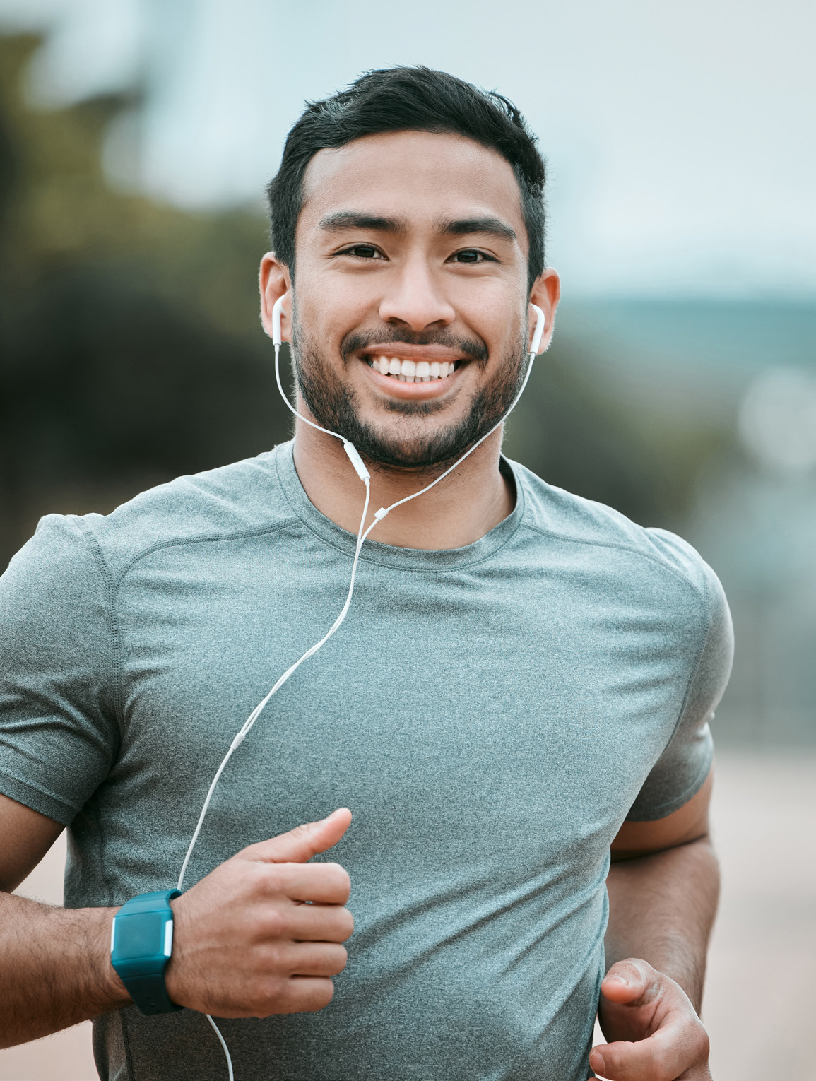 Smiling young man in a gray athletic shirt wearing white earbuds, running outdoors with a blurry background. He has a blue fitness watch on his wrist and is wearing a short-sleeved moisture-wicking shirt, looking happy and energetic during his workout.